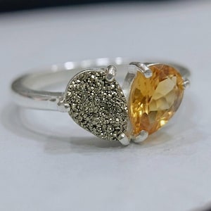 Op de afbeelding: Een zilveren ring met twee naast elkaar geplaatste druppelvormige stenen. De ene steen is een getextureerde, metallic grijze druzy, en de andere is een gefacetteerde, doorschijnende gele citrien. De stenen zitten vast met kleine zilveren pootjes.