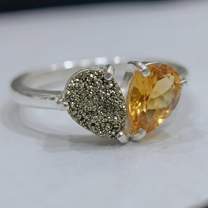 Op de afbeelding: Een zilveren ring met twee naast elkaar geplaatste druppelvormige stenen. De ene steen is een getextureerde, metallic grijze druzy, en de andere is een gefacetteerde, doorschijnende gele citrien. De stenen zitten vast met kleine zilveren pootjes.
