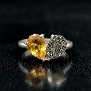 Op de afbeelding: Een zilveren ring met twee peervormige stenen die naast elkaar een hartvorm vormen. De ene steen is een gefacetteerde, doorschijnende gele citrien, de andere is een getextureerde, metallic grijze pyriet met een sprankelend oppervlak.