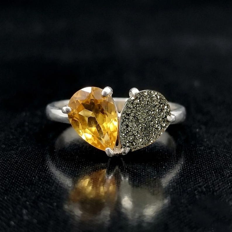 Op de afbeelding: Een zilveren ring met twee peervormige stenen die naast elkaar een hartvorm vormen. De ene steen is een gefacetteerde, doorschijnende gele citrien, de andere is een getextureerde, metallic grijze pyriet met een sprankelend oppervlak.