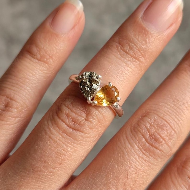 Op de afbeelding: Een zilveren ring met twee stenen naast elkaar. De ene steen is een getextureerde, metallic pyriet met een kubusvormige kristalstructuur, en de andere is een gladde, doorschijnende, peervormige gele citrien gezet in een zilveren band.