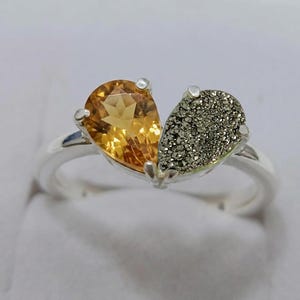 Op de afbeelding: Een zilveren ring met twee peervormige stenen naast elkaar. De ene steen is een gefacetteerde, doorschijnende goudgele citrien, terwijl de andere een getextureerde, metaalgrijze pyriet is. De stenen worden door zilveren klauwen op de ring gehouden.