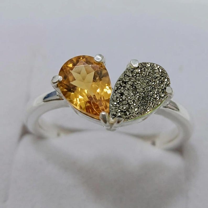 Op de afbeelding: Een zilveren ring met twee peervormige stenen naast elkaar. De ene steen is een gefacetteerde, doorschijnende goudgele citrien, terwijl de andere een getextureerde, metaalgrijze pyriet is. De stenen worden door zilveren klauwen op de ring gehouden.