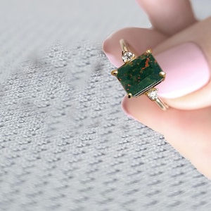 Puede incluir: Un anillo de oro con una piedra preciosa verde oscura de talla esmeralda y dos pequeños diamantes transparentes en la banda.