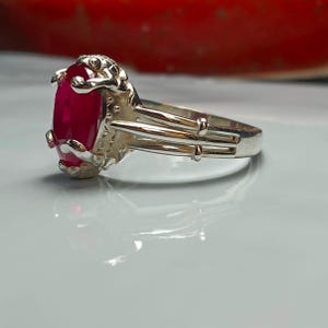 Puede incluir: Un anillo de plata con una gran piedra preciosa de rubí de forma ovalada engastada en una garra. La banda del anillo tiene tres bandas decorativas.