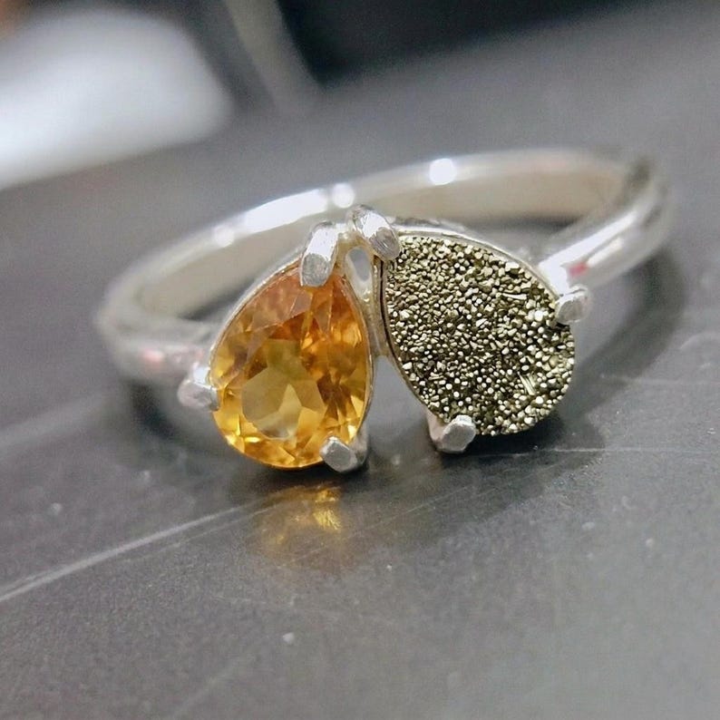 Op de afbeelding: Een zilveren ring met twee peervormige stenen naast elkaar. De ene steen is een gefacetteerde, doorschijnende gele citrien en de andere is een getextureerde, metaalachtig uitziende pyriet druzy. De ringband is van glad, gepolijst zilver.