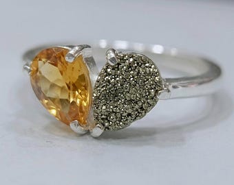 May include: A silver ring featuring two side-by-side pear-shaped stones. One stone is a faceted, translucent golden-yellow citrine, while the other is a textured, metallic gray pyrite druzy. Both stones are held in place by silver prongs.