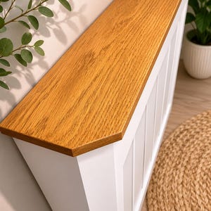 May include: A radiator cover featuring a white vertical slatted base and a thick, natural oak-coloured wooden top shelf. The structure has a rectangular design with clean lines, providing a flat surface area above the white vertical paneling.