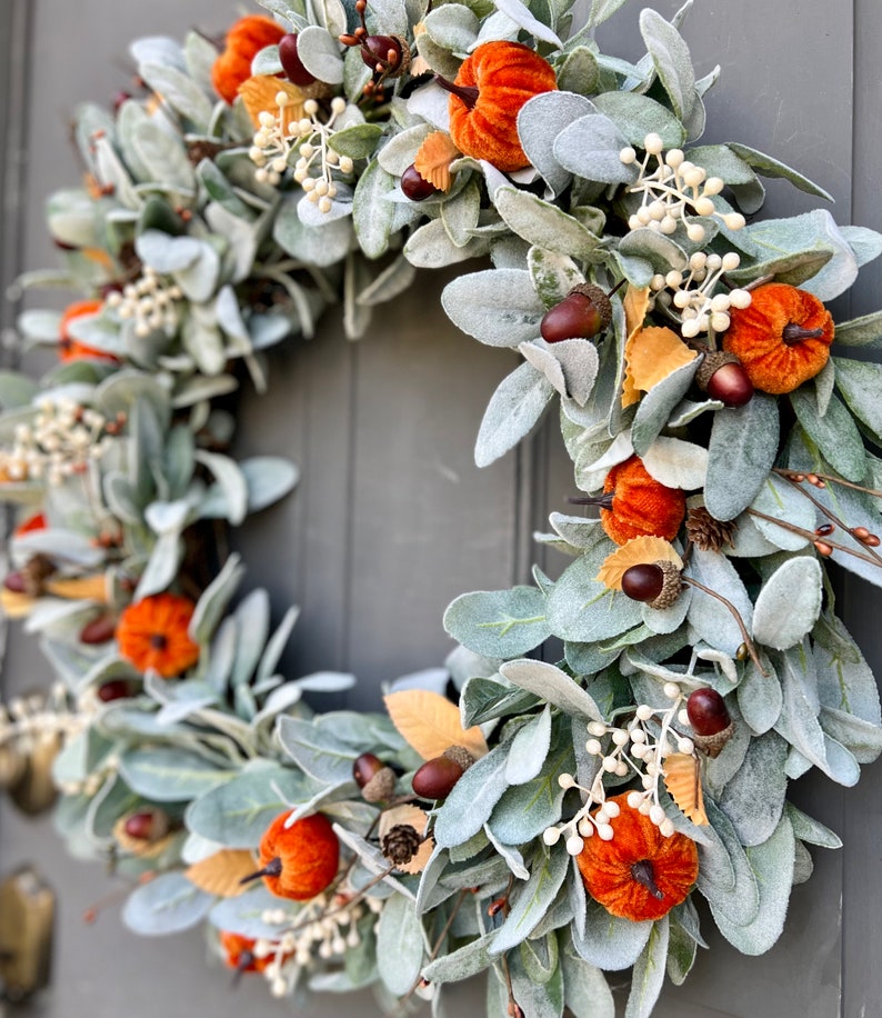 Puede incluir: Una corona de oto&ntilde;o con base de hojas verdes de oreja de cordero, adornada con calabazas de terciopelo en tonos naranja y marr&oacute;n, bellotas y bayas blancas.