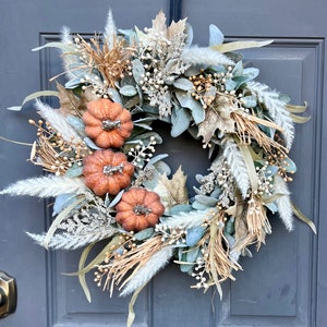 May include: A fall-themed wreath with a base of green eucalyptus leaves, accented with dried grasses, white pampas grass, and small faux pumpkins.