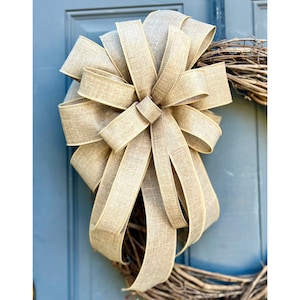 May include: A large, decorative bow made of tan burlap ribbon. The bow is attached to a grapevine wreath, with the bow taking up most of the frame. The background is a blue door.