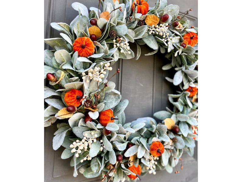 Puede incluir: Una corona de oto&ntilde;o con base de hojas de oreja de cordero verdes y blancas. La corona est&aacute; decorada con peque&ntilde;as calabazas de terciopelo naranja, bellotas marrones y bayas blancas.