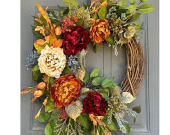 May include: A decorative wreath featuring a natural grapevine base adorned with artificial flowers in shades of deep burgundy, burnt orange, and cream. The arrangement includes green foliage, small blue berries, and textured stems, creating a full, layered display of autumn-inspired colors.