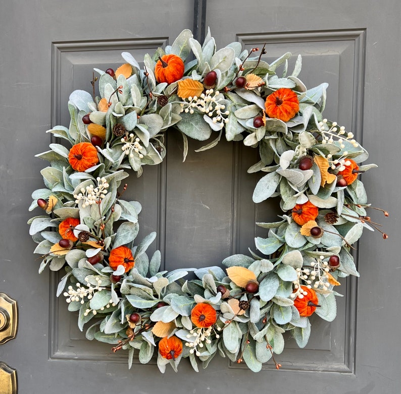 Puede incluir: Una corona de oto&ntilde;o con una base de follaje seco verde plateado. La corona est&aacute; decorada con peque&ntilde;as calabazas de fieltro naranja y bayas blancas.
