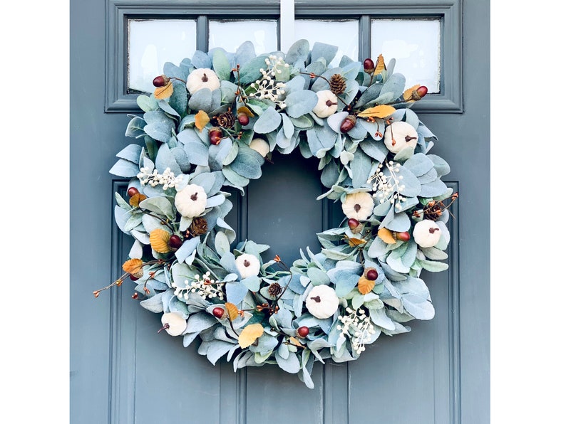 Puede incluir: Una corona azul y blanca con peque&ntilde;as calabazas blancas y detalles marrones. La corona est&aacute; hecha de hojas de eucalipto y es perfecta para la decoraci&oacute;n de oto&ntilde;o.