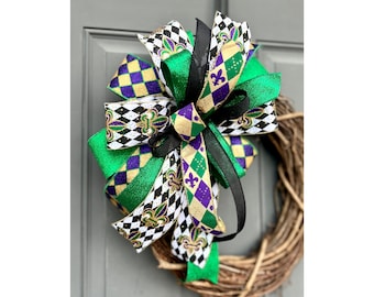 May include: A decorative wreath bow featuring a vibrant mix of colors and patterns. The bow incorporates green, purple, black, and gold ribbons with harlequin and fleur-de-lis designs. The bow is attached to a natural grapevine wreath.