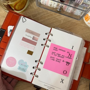 May include: An open, orange leather planner with white pages and a red spine. The planner contains sticky notes, stickers, and handwritten notes. A pink sticky note reads "check notebooks for paper, add more pics -> photos to my planner, print inserts for ing. (stats check)".