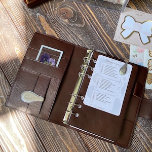Op de afbeelding: Open bruin leren planner met zilveren bindringen. Een kaart met "Planning routines" is zichtbaar, samen met een kleine kaart met geperste bloemen. De planner ligt open op een houten oppervlak, met briefpapier en stickers op de achtergrond.
