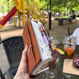 Op de afbeelding: Een lichtbruine lederen planner met een metalen sluiting, met witte pagina's en tabbladen met de labels Month, Today, To Do List en Movies. Een paarse en zilveren pen is aan de zijkant van de planner bevestigd, die is voorzien van een voorvak.