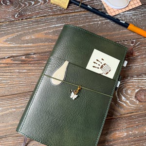 Może przedstawiać: Zielony skórzany notes z białą zawieszką w kształcie motyla przymocowaną do sznurka. Notes ma białą skórzaną kieszeń z przodu z małą białą naklejką z napisem "Cappuccino" w języku japońskim. Notes leży na drewnianej powierzchni.