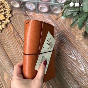 May include: A tan leather journal with a dark brown elastic closure. A tag with a handprint and a QR code is attached. The journal is held against a wooden surface, with washi tape rolls and greenery in the background.