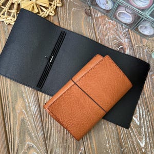 May include: A black leather journal cover with an open design, next to a closed, textured brown leather journal. The journals are on a wooden surface. Decorative washi tape rolls are visible in the background.