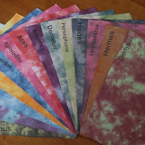 Puede incluir: Un conjunto de 12 cuadrados de tela con patrones tie-dye en varios colores. Cada cuadrado tiene escrito el nombre de un dios o diosa griego, incluyendo Zeus, Hera, Atenea, Apolo, Poseidón, Afrodita, Ares, Artemisa, Deméter, Perséfone, Hades, Hefesto, Hermes y Dionisio.