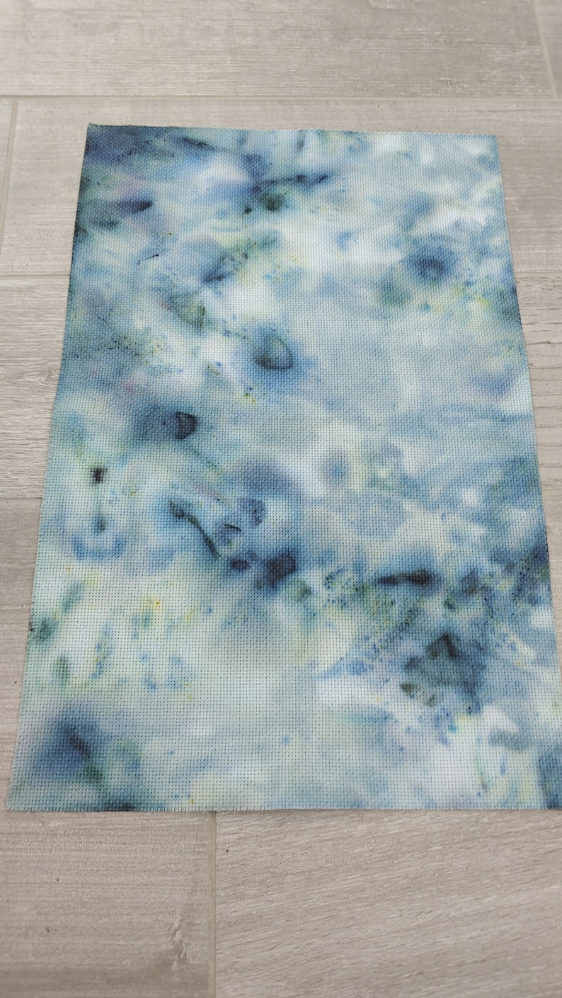 May include: A piece of white fabric with a blue and green tie-dye pattern. The fabric has a subtle grid pattern.