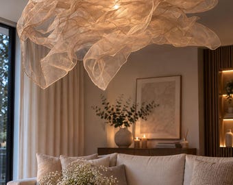 May include: A large, sculptural pendant light fixture featuring layers of translucent, beige mesh fabric that create a cloud-like, organic form. The light is suspended by a thin cord and emits a warm, soft glow from a central light source.
