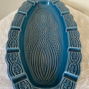 Peut inclure: Un cendrier en céramique bleu de forme ovale avec un motif décoratif. Le cendrier a un bord surélevé avec un motif répétitif d'étoiles et de cercles. Le centre du cendrier présente un motif géométrique.