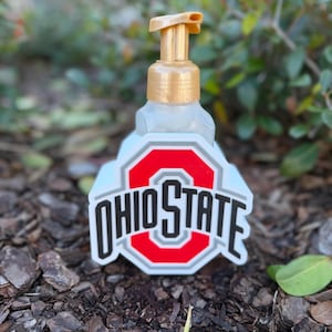 Könnte beinhalten: Ein klarer Seifenspender mit goldener Pumpe und dem Ohio State-Logo in Rot, Weiß und Schwarz. Der Spender steht auf einer Mulchschicht mit grünem Laub im Hintergrund.