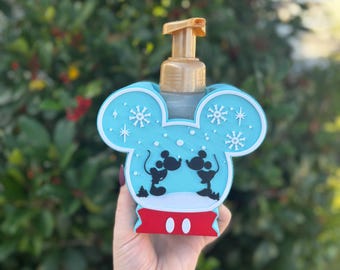 May include: A light blue Mickey Mouse-shaped soap dispenser with a gold pump. The dispenser features a winter scene with Mickey and Minnie Mouse silhouettes and snowflake details. The base is red with two white dots.