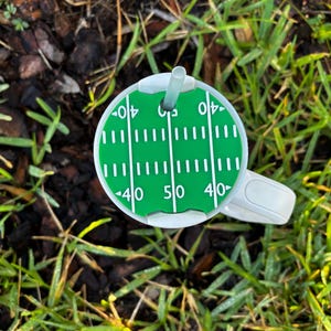 May include: A white and green football field-themed drinks holder with a straw. The drink holder features yard lines and the numbers 40 and 50. The drink holder is sitting on grass and mulch.