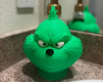 Puede incluir: Un dispensador de jabón verde con temática de Grinch con una bomba dorada. El dispensador tiene una cara esculpida con ojos blancos y una nariz negra. El dispensador está sobre una encimera.