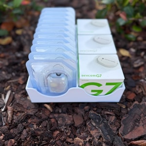 May include: A white tray contains multiple Dexcom G7 medical devices. The tray holds clear plastic packaging with medical devices and white boxes with the Dexcom G7 logo. The background is blurred, with brown mulch and green foliage.