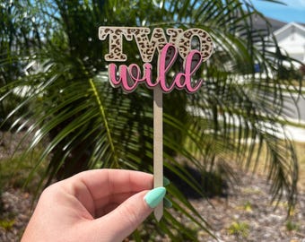 May include: A wooden cake topper with the words "TWO wild" in pink and brown leopard print. The topper is on a wooden stick.