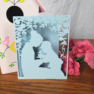 May include: A light blue paper cut-out card featuring a mother and child silhouette under a floral arch. The card is on a wooden surface, with a pink birdhouse and paper roses in the background.