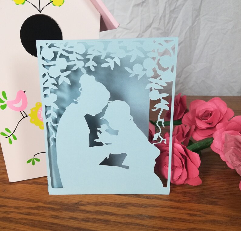 May include: A light blue paper cut-out card featuring a mother and child silhouette under a floral arch. The card is on a wooden surface, with a pink birdhouse and paper roses in the background.