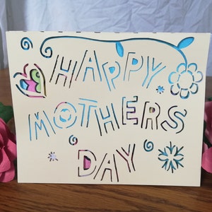 May include: A cream-colored Mother's Day card with cut-out lettering that reads "HAPPY MOTHERS DAY". The card features floral and heart-shaped cutouts with colorful accents. Two pink paper roses are on the right.