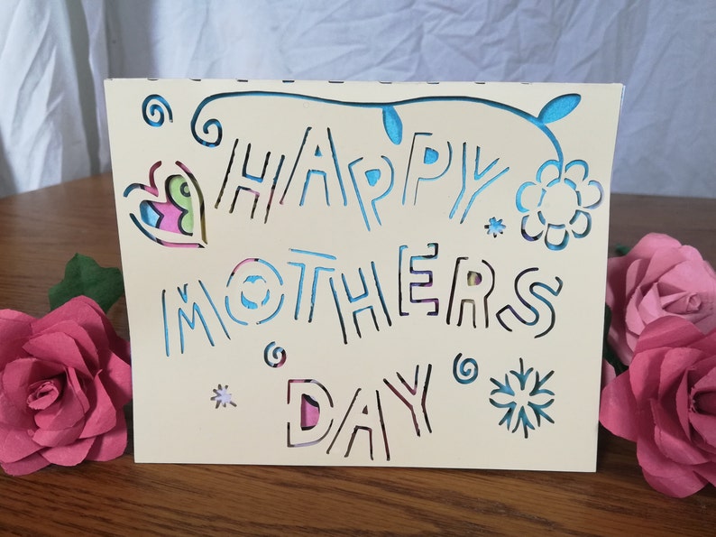 May include: A cream-colored Mother's Day card with cut-out lettering that reads "HAPPY MOTHERS DAY". The card features floral and heart-shaped cutouts with colorful accents. Two pink paper roses are on the right.