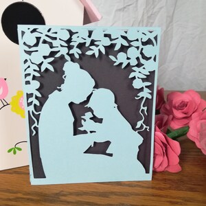 May include: A light blue paper cut-out card featuring a mother and child under a floral arch. The card has a dark gray background and is surrounded by pink paper roses and a decorative birdhouse.