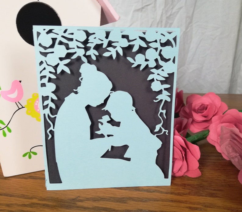 May include: A light blue paper cut-out card featuring a mother and child under a floral arch. The card has a dark gray background and is surrounded by pink paper roses and a decorative birdhouse.