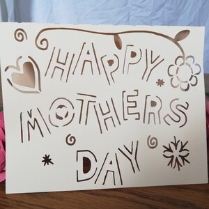 May include: Cream-colored Mother's Day card with the words "HAPPY MOTHERS DAY" cut out in a whimsical font. The card features decorative cut-out hearts, flowers, and swirls. A pink paper rose is visible in the background.