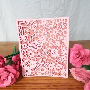 May include: A light pink, laser-cut paper card with a floral design. The card features intricate cutouts of flowers, leaves, and a butterfly. The card is displayed on a wooden surface, with pink paper roses in the foreground.