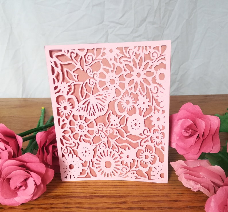 May include: A light pink, laser-cut paper card with a floral design. The card features intricate cutouts of flowers, leaves, and a butterfly. The card is displayed on a wooden surface, with pink paper roses in the foreground.