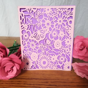 May include: A light pink, laser-cut floral design card with a purple backing. The card features intricate cut-out patterns of flowers and leaves. Pink paper roses are arranged on a wooden surface, enhancing the card's delicate aesthetic.