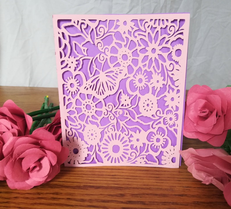 May include: A light pink, laser-cut floral design card with a purple backing. The card features intricate cut-out patterns of flowers and leaves. Pink paper roses are arranged on a wooden surface, enhancing the card's delicate aesthetic.