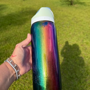 May include: A cylindrical water bottle with a vibrant, metallic rainbow gradient finish transitioning from deep blue to green, yellow, and magenta. The bottle features a white lid with the brand name Owala embossed on the front and a light blue accent.