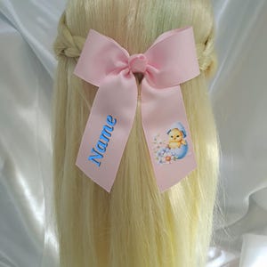 May include: A pale pink hair bow with the word "Name" in blue script on one tail. The other tail features a cartoon chick hatching from a blue egg with floral accents. The bow is attached to blonde hair.