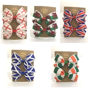 May include: Set of two hair bows featuring different national flags. The bows are made of white ribbon with printed flags of England, Wales, Scotland, Ireland, and the United Kingdom. Each bow is attached to a brown card with the text "Thank you".